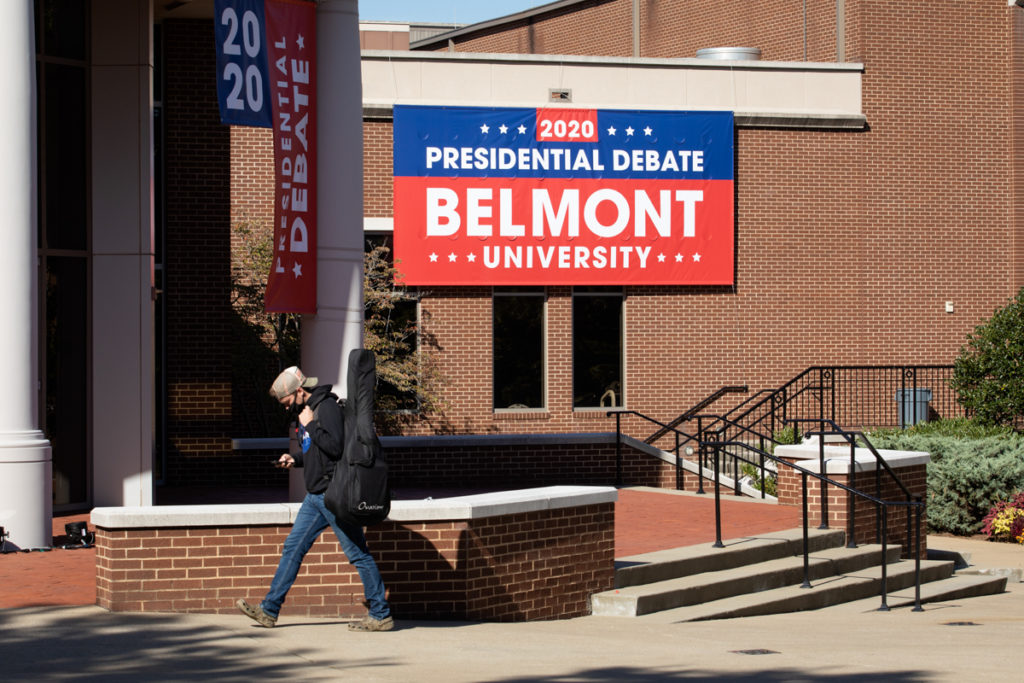 A person in a mask walks past a sign announcing the Presidential Debate at Belmont University.