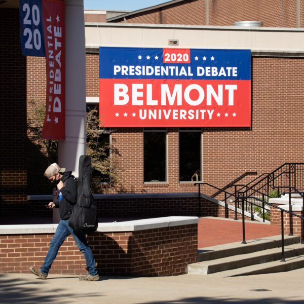 A person in a mask walks past a sign announcing the Presidential Debate at Belmont University.