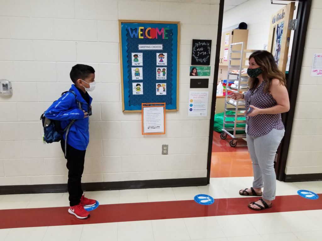 A teacher greets a student while wearing masks in a school building.