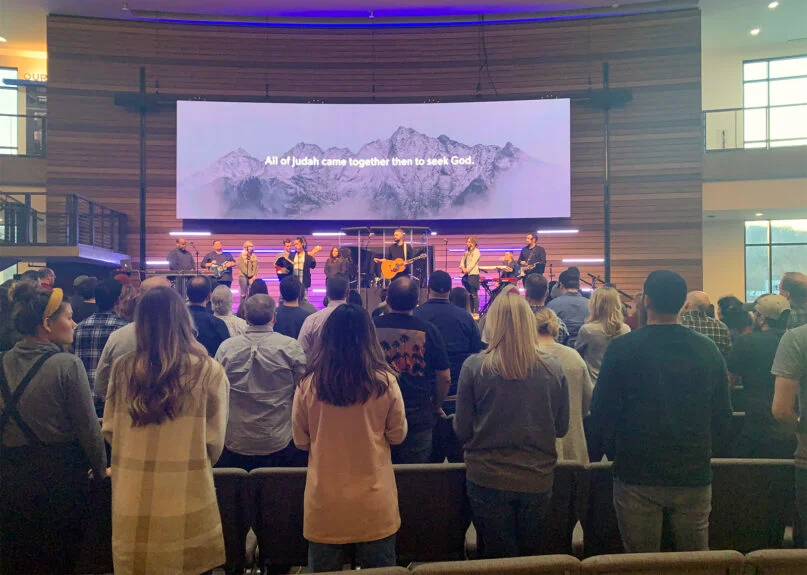 Praise and Worship service seen from behind the crowd, with a band on stage and a screen with religious text behind the band