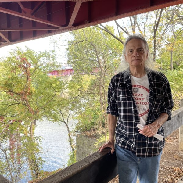 Native American historian Toye Heape, wearing a plaid shirt and a Native History Association T-shirt, stands in front of a railing; in the near distance, there is stone structure that was once part of a bridge.