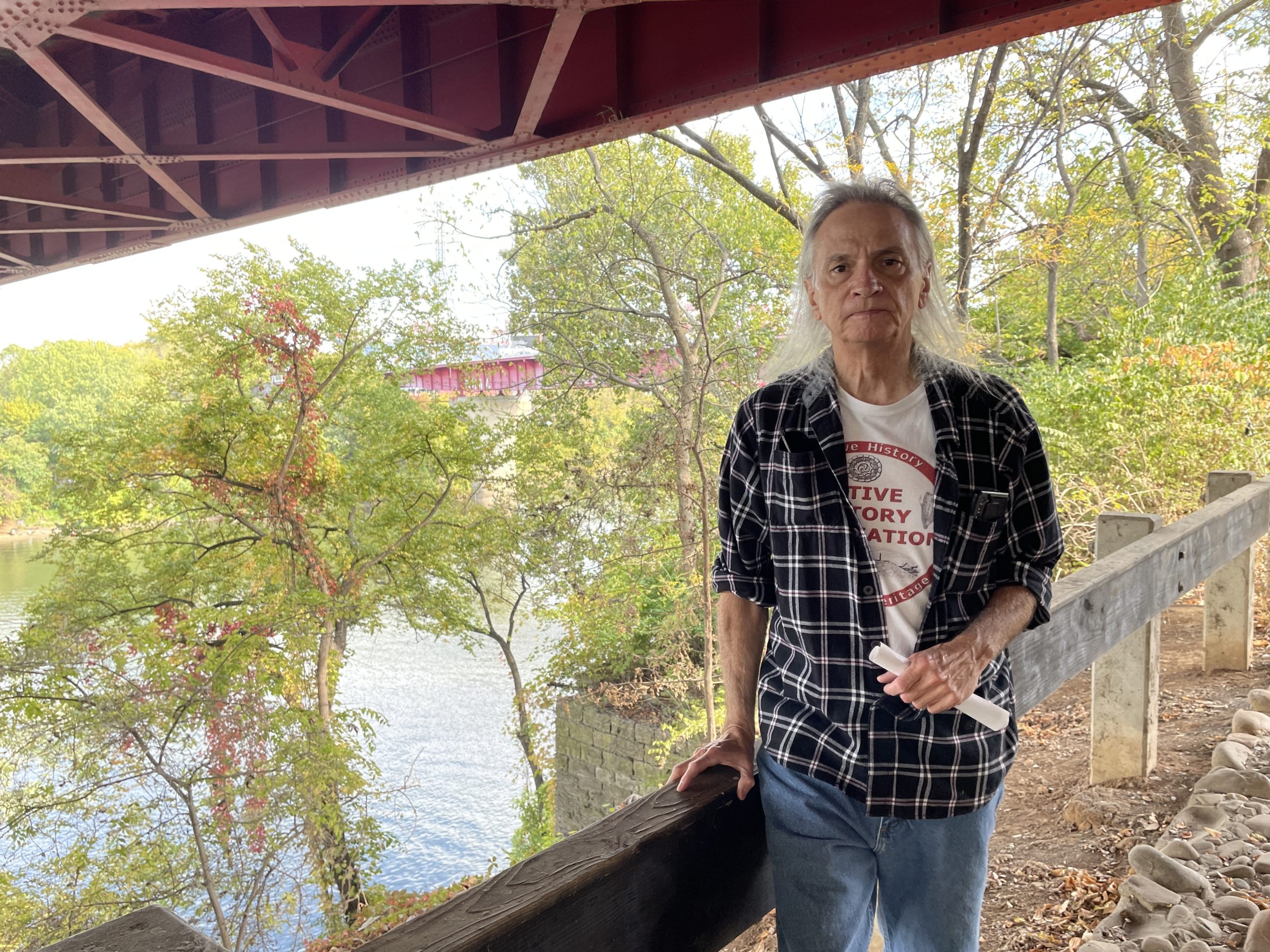 Native American historian Toye Heape, wearing a plaid shirt and a Native History Association T-shirt, stands in front of a railing; in the near distance, there is stone structure that was once part of a bridge.