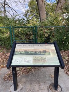 An interpretive plaque in front of a fence; the plaque reads: "Journey West: Forced Removal of the Cherokee" and includes a drawing of the bridge that once stood here, spanning the Cumberland River.