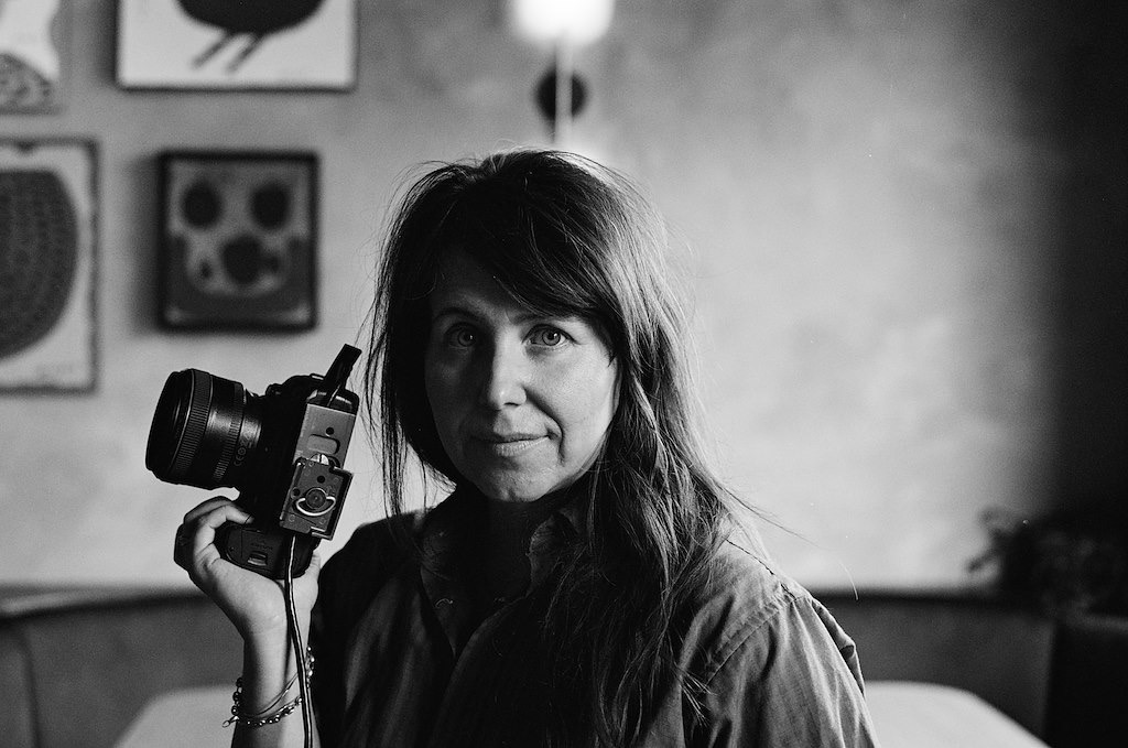 In a black and white photo, photographer Emily Dorio, a white woman, looks at the camera while holding a camera in her right hand