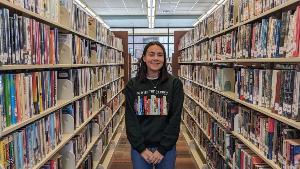 Julia Garnett, 18, smiles standing among the book shelves at Hendersonville Public Library. Her shirt, decorated with the spines of books that have been challenged across the country, reads "I'm with the banned."