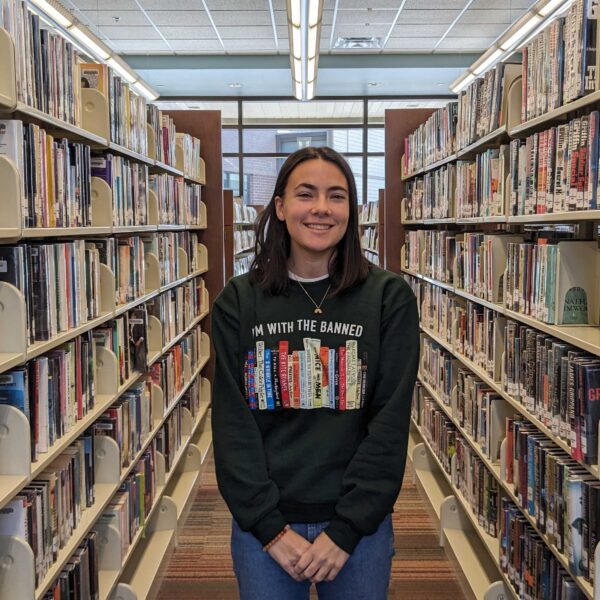 Julia Garnett, 18, smiles standing among the book shelves at Hendersonville Public Library. Her shirt, decorated with the spines of books that have been challenged across the country, reads "I'm with the banned."