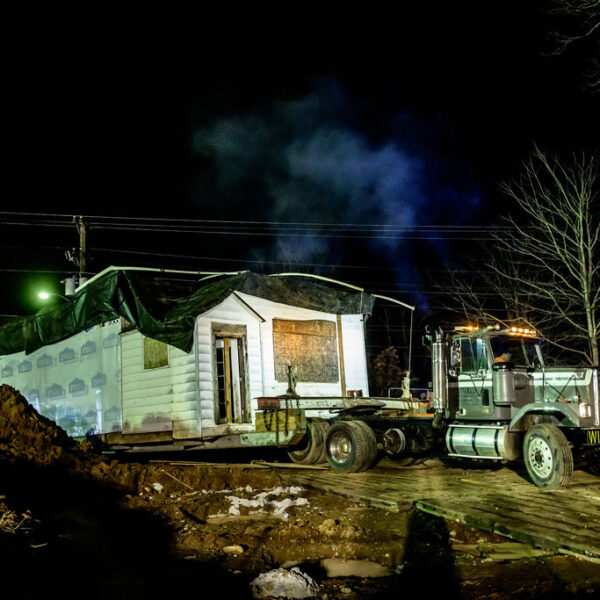 A white clapboard schoolhouse is unloaded from the back of a semi-truck, in darkness. The ground below the truck is a pile of dirt.