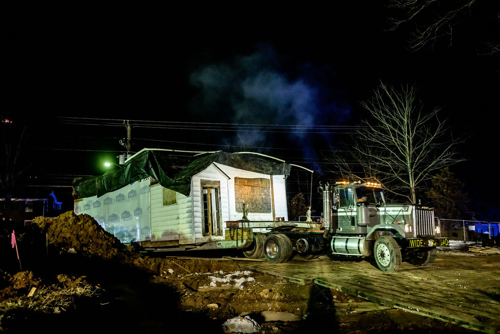 A white clapboard schoolhouse is unloaded from the back of a semi-truck, in darkness. The ground below the truck is a pile of dirt.