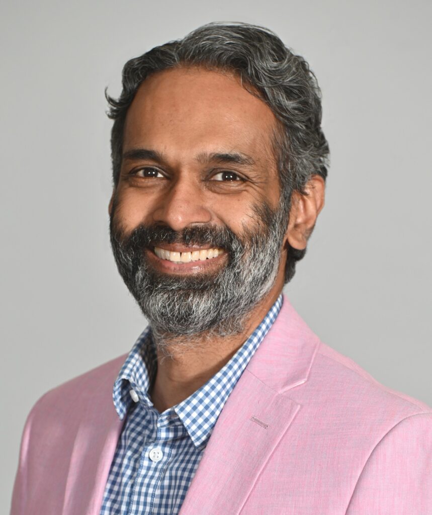 Daniel Singh, a man with grayish-black hair and a beard, wearing a pink suit jacket, smiles at the camera in front of a white background