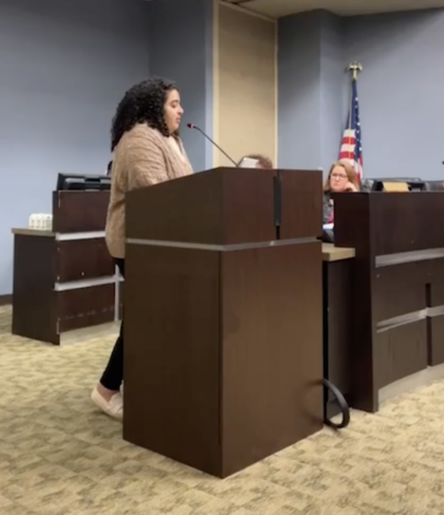 Lydia Yousief, a woman with black curly hair and wearing a light brown sweater, speaks at a podium in a meeting room. A commissioner is seated at the table behind her.