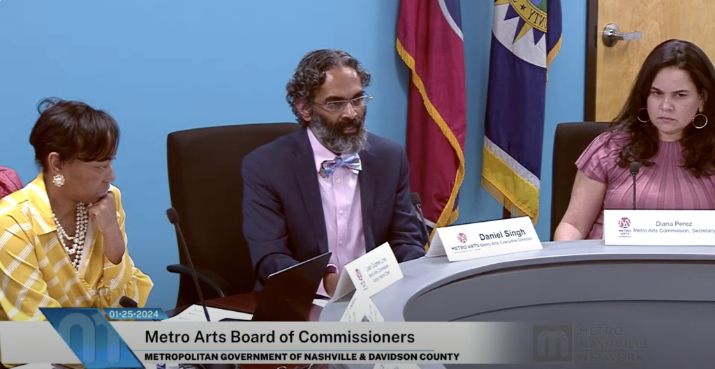 Metro Arts Commissioner Leah Dupree Love, Director Daniel Singh, and Commissioner Diana Perez sit in office chairs at a boardroom table in front of a blue wall. The Tennessee flag is on a pole behind them.