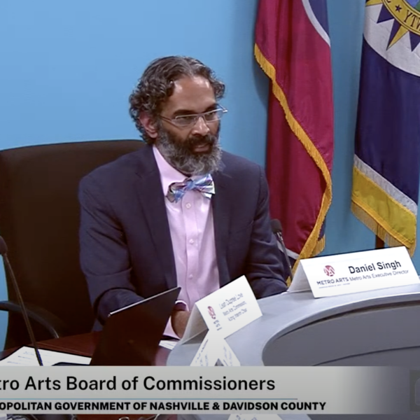 Metro Arts Commissioner Leah Dupree Love, Director Daniel Singh, and Commissioner Diana Perez sit in office chairs at a boardroom table in front of a blue wall. The Tennessee flag is on a pole behind them.