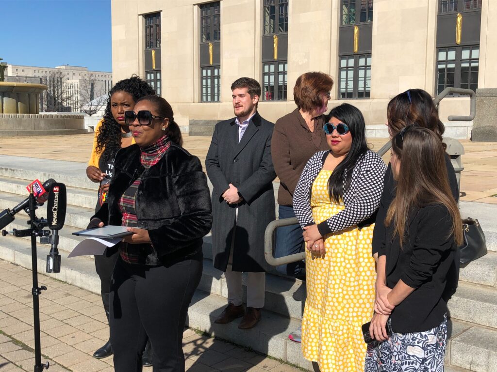 A woman with sunglasses stands at a microphone and several people stand behind her. They are wearing coats. In the background are stairs, an art-deco courthouse, and a bright blue sky.