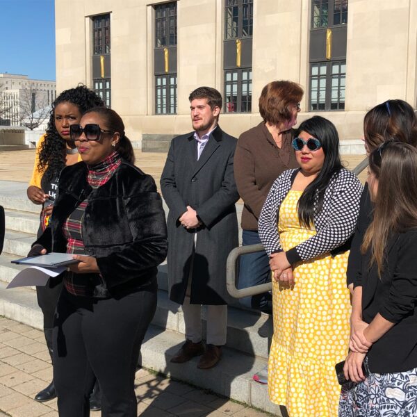 A woman with sunglasses stands at a microphone and several people stand behind her. They are wearing coats. In the background are stairs, an art-deco courthouse, and a bright blue sky.