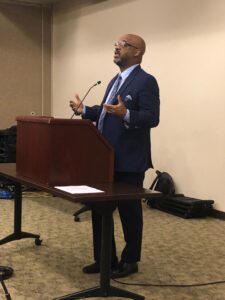 A bald man with glasses and a trimmed beard stands at a lectern in a meeting room. He wears a blue suit and tie.