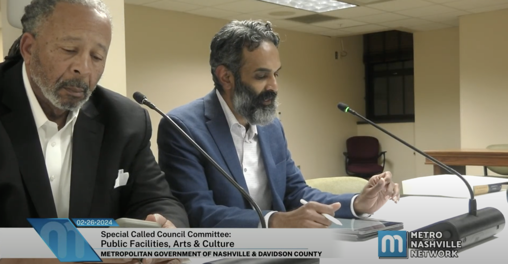 Davie Tucker, a tall man with a trimmed beard and gray dreadlocks, and Daniel Singh, a man with wavy hair and a graying black beard, sit next to each other in a meeting room and speak into microphones. Both men are wearing suits.