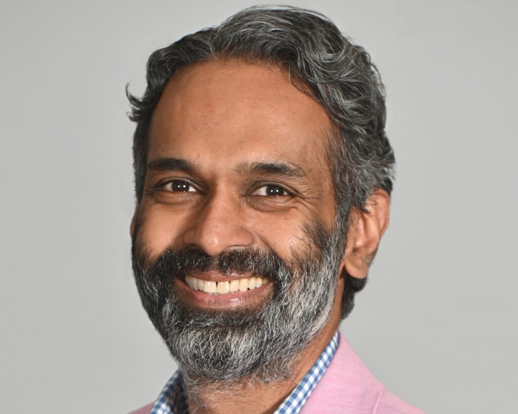 Daniel Singh, a man with grayish-black hair and a beard, wearing a pink suit jacket, smiles at the camera in front of a white background