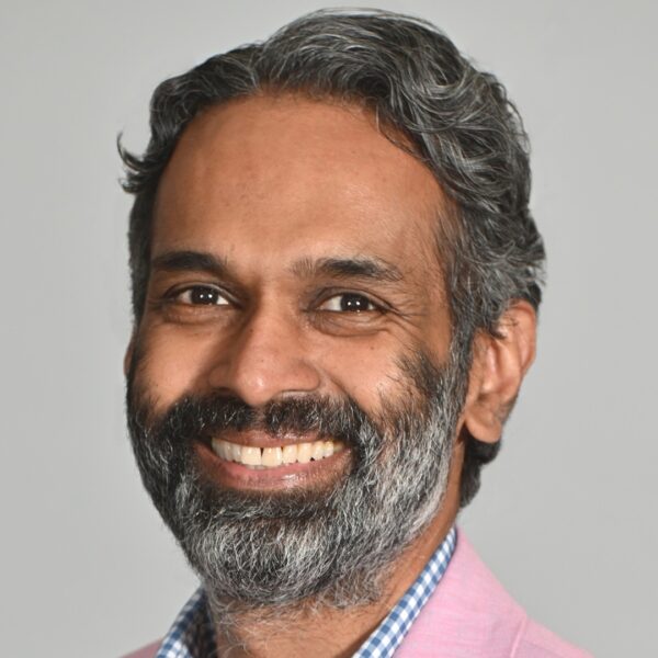 Daniel Singh, a man with grayish-black hair and a beard, wearing a pink suit jacket, smiles at the camera in front of a white background