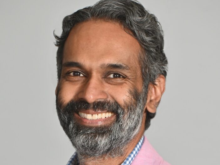 Daniel Singh, a man with grayish-black hair and a beard, wearing a pink suit jacket, smiles at the camera in front of a white background