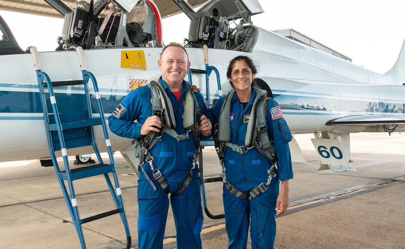 Two astronauts in flight suits stand in front of a fighter jet smiling for the camera.