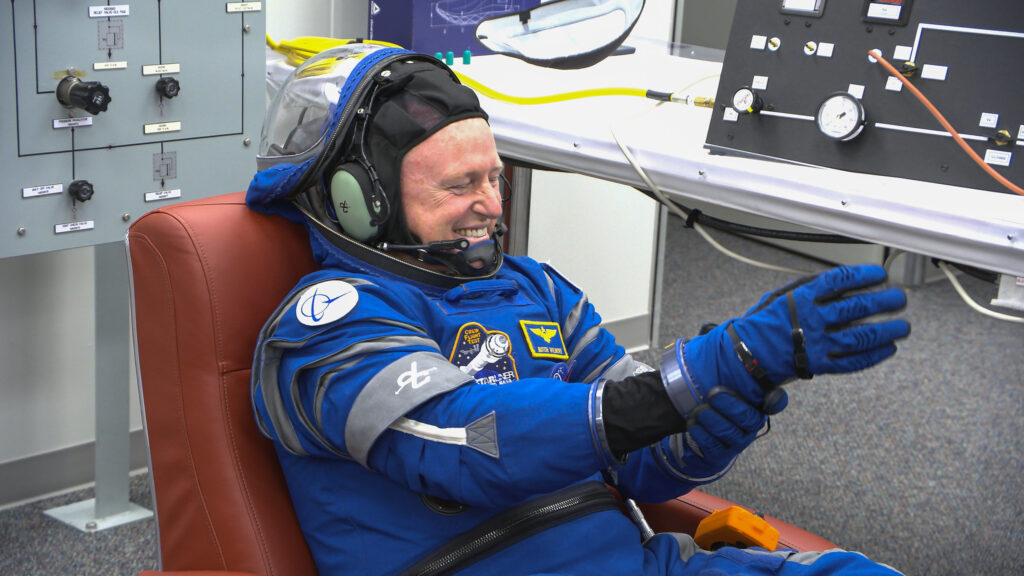 An astronaut in a blue spacesuit smiles while sitting in a chair