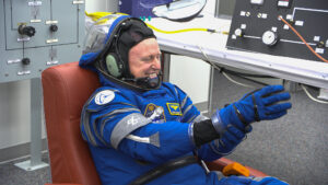 An astronaut in a blue spacesuit smiles while sitting in a chair