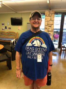 A man with a baseball cap, pink shorts, a water bottle, a nametag, and a blue t-shirt with a rainbow, a unicorn head and the following text: "Drag queens are fabulous & magical like unicorns only better"