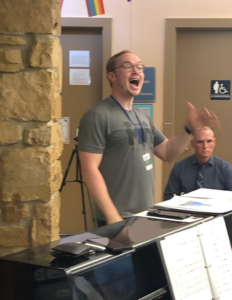 A man with blond hair, glasses and a green t-shirt conducts a choir with his left hand. His face is expressive as he mouths the words the choir sings.