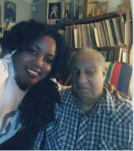 Two people: a woman with long black hair and an elderly blind man in a checkered blue button-down shirt, pose for a selfie in front of a bookcase.