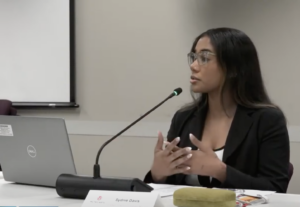 A woman with long black hair wearing a black pantsuit talks into a microphone while seated at a table in a meeting room.