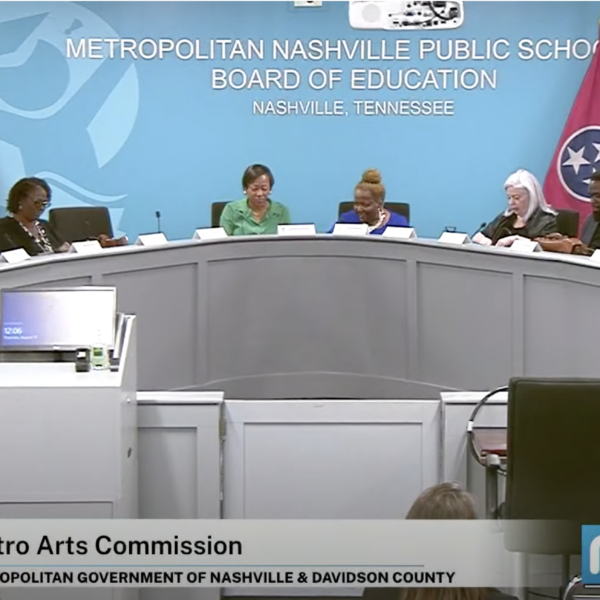 A group of public officials in formal attire sit at a dais, with a blue wall and a Tennessee flag behind them.