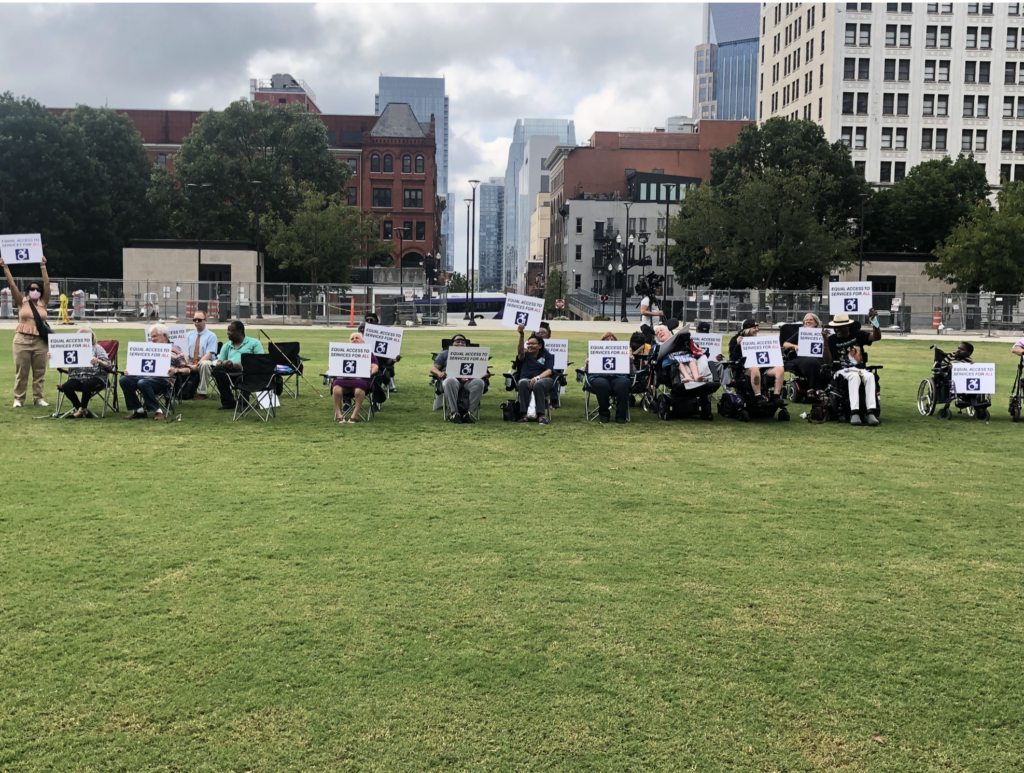 A group of people, some in wheelchairs, sit in folding chairs on a lawn with tall buildings behind them. Some hold up protest signs.