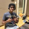 A teenage boy wearing a t-shirt, glasses and headphones smiles and gives two thumbs up. He sits in front of an orange microphone in a radio studio.