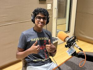 A teenage boy wearing a t-shirt, glasses and headphones smiles and gives two thumbs up. He sits in front of an orange microphone in a radio studio.