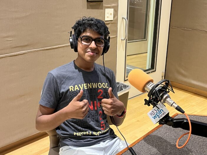 A teenage boy wearing a t-shirt, glasses and headphones smiles and gives two thumbs up. He sits in front of an orange microphone in a radio studio.