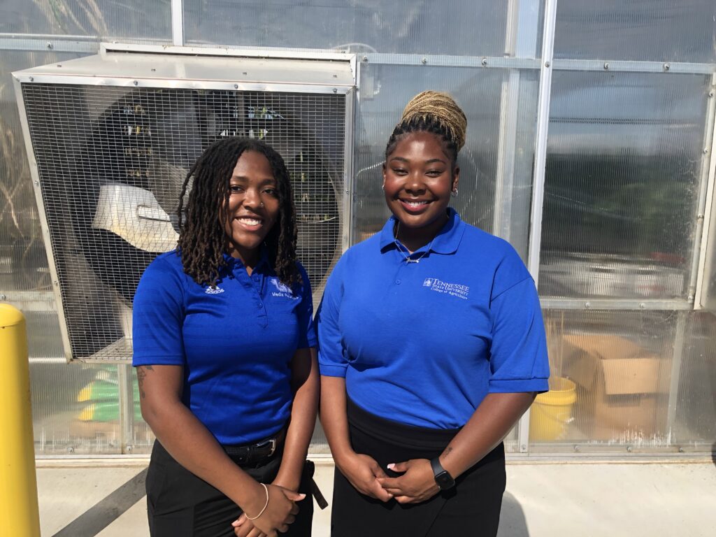 Two women in blue polo shirts pose for a photo in front of a greenhouse