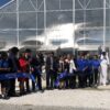 A group of well-dressed people cut a ribbon in front of a greenhouse on a sunny day. The man in the center carries a pair of giant scissors.