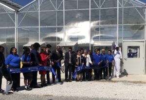 A group of well-dressed people cut a ribbon in front of a greenhouse on a sunny day. The man in the center carries a pair of giant scissors.