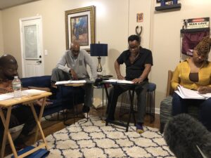 Four actors sit on couches and armchairs and read scripts set in front of them on tables in a warmly-lit living room.