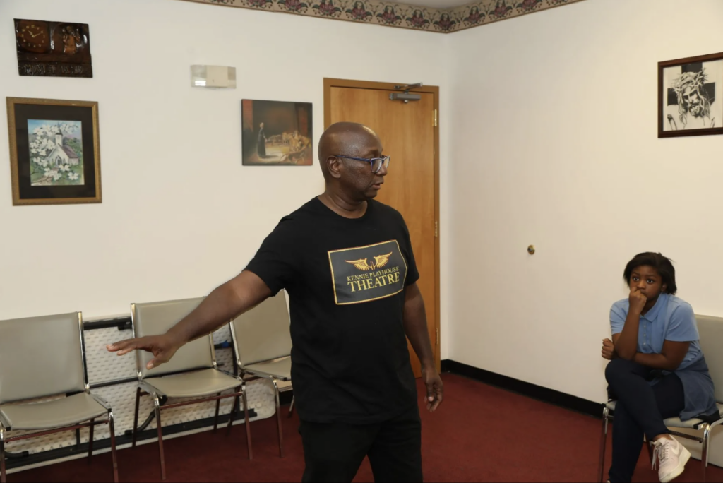 A director talks and points with his finger in a carpeted rehearsal room. A few chairs are set up behind him.