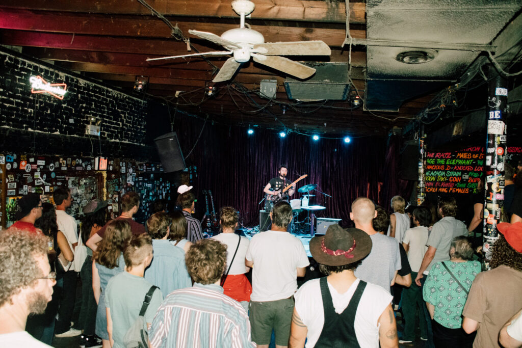 A packed concert venue with wood beams on the ceiling and a bar. A guitarist plays on stage.