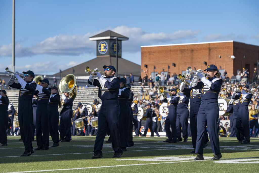 Trumpeters in a marching band hold up their instruments and play in formation. They wear blue uniforms and stand on a football field under a clear blue sky.