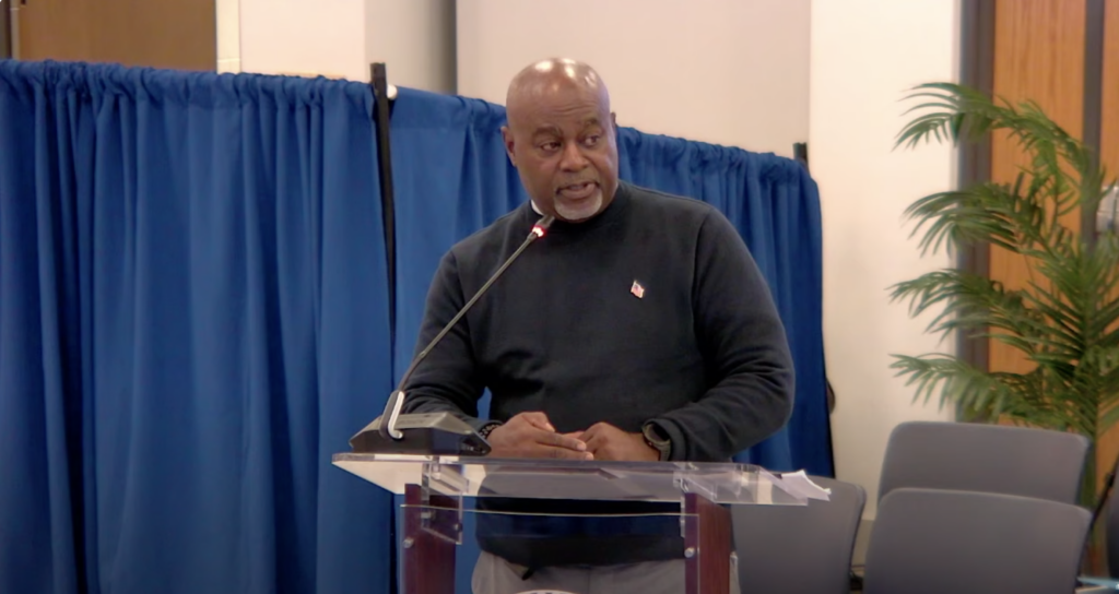 A man stands at a lectern in front of a blue curtain and a tall fern.