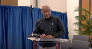 A man stands at a lectern in front of a blue curtain and a tall fern.