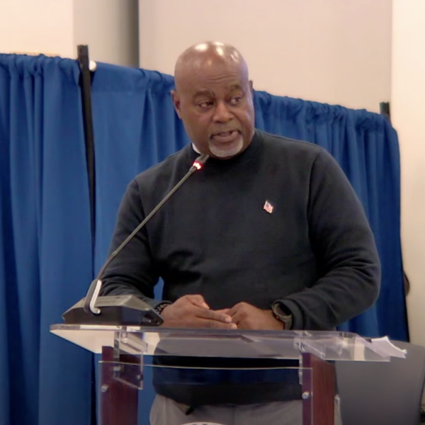 A man stands at a lectern in front of a blue curtain and a tall fern.