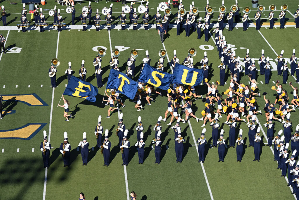 Looking down on a marching band marching in a square formation on a football field. in the middle, band members hold up four flags, each with a letter: "E," "T," "S," and "U."