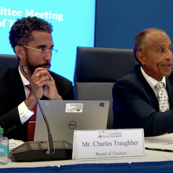 Two men in suits sit at a table, behind nameplates and in front of a large blue television screen.