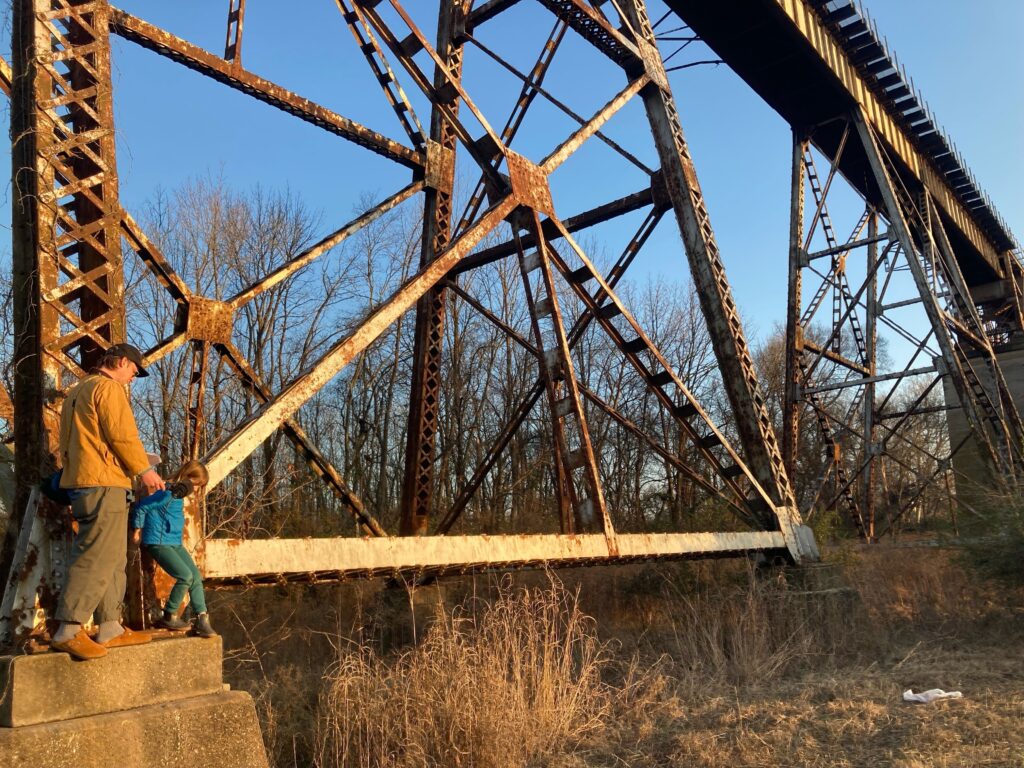 An adult and a kid on in golden hour light standing under a large train bridge