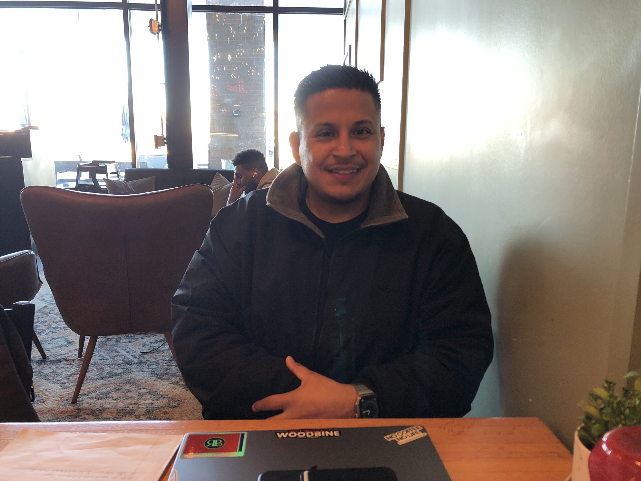 A man in a blue coat sits at a table in a coffee shop. Light shines in from a glass wall behind him.