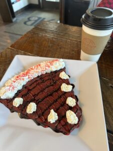 A red crepe folded into a triangle with dots of whipped cream. A paper coffee cup sits behind it on a wooden table.